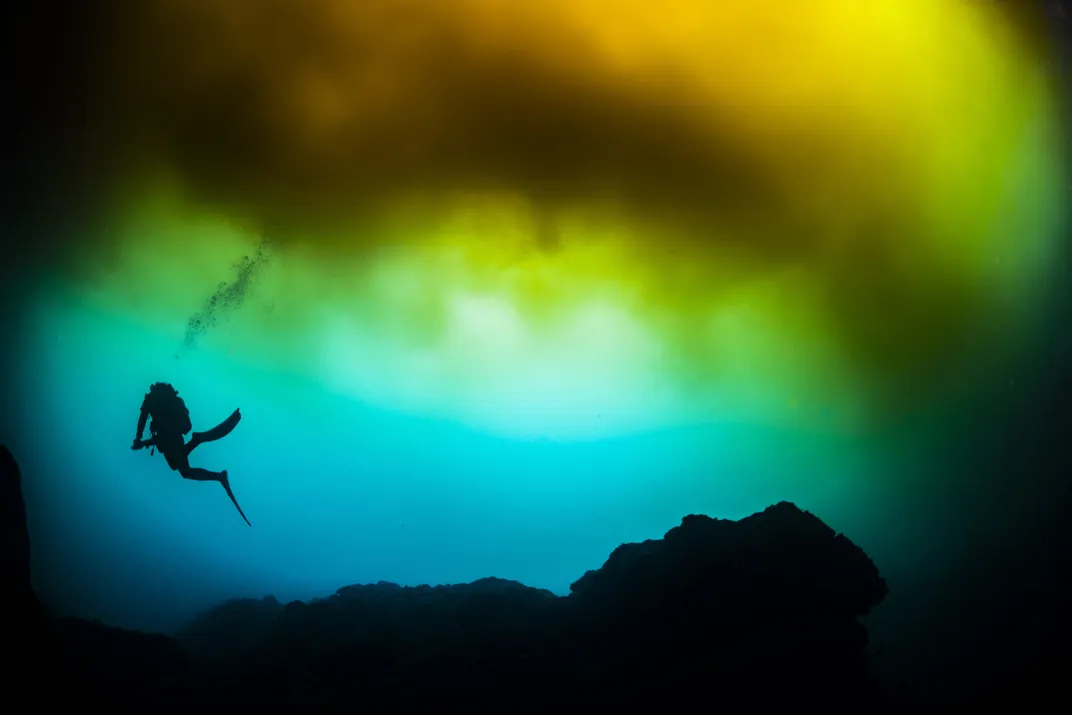 A diver swimming through blue, red, orange, and green water colored by a hot spring.