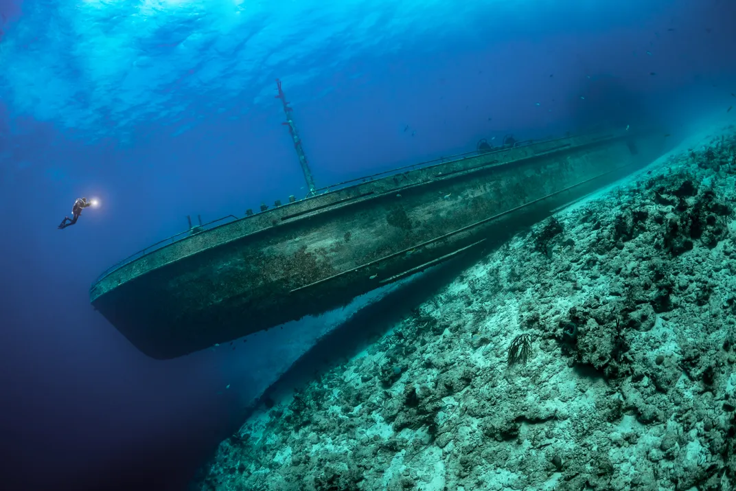 A scuba diver next to a large shipwreck on top of a rocky ocean floor.