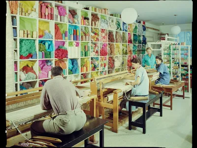 A smiling Dorothy Liebes stands in her studio in her blue apron, supervising two young women sitting at looms with spools of colorful thread and skeins of yarn close at hand.