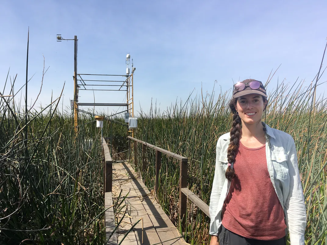 Ariane Arias-Ortiz, a young light-skinned female scientist with a long brown braid, stands in a wetland on a wooden boardwalk, with a research tower behind her.