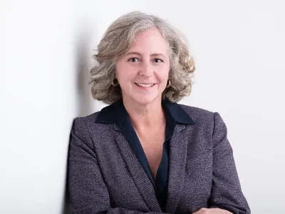 Elizabeth Babcock from the waist up against a white background. She is leaning against a wall with her arms crossed in front of herself.