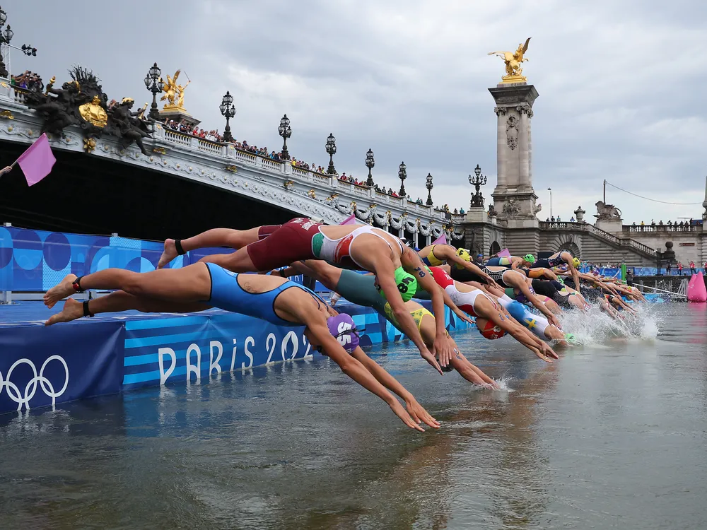 A line of athletes diving into a river with a bridge in the background