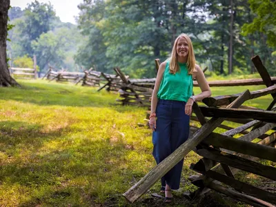 A woman leans on a snaking, split rail fence on a mossy green field.