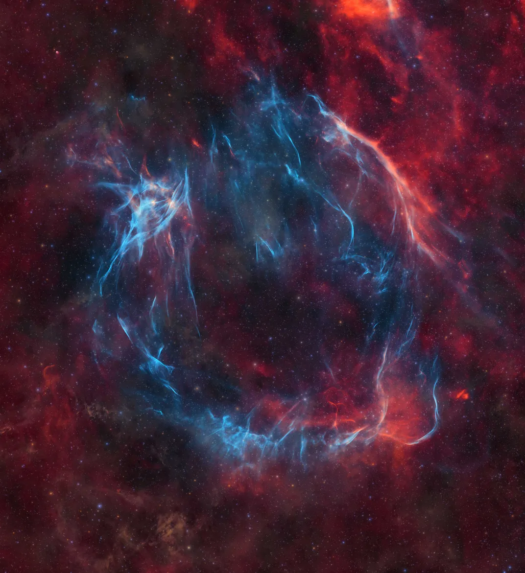 Bright blue remnants of a gigantic supernova form a wispy ring against red remnants and a red foggy backdrop with stars